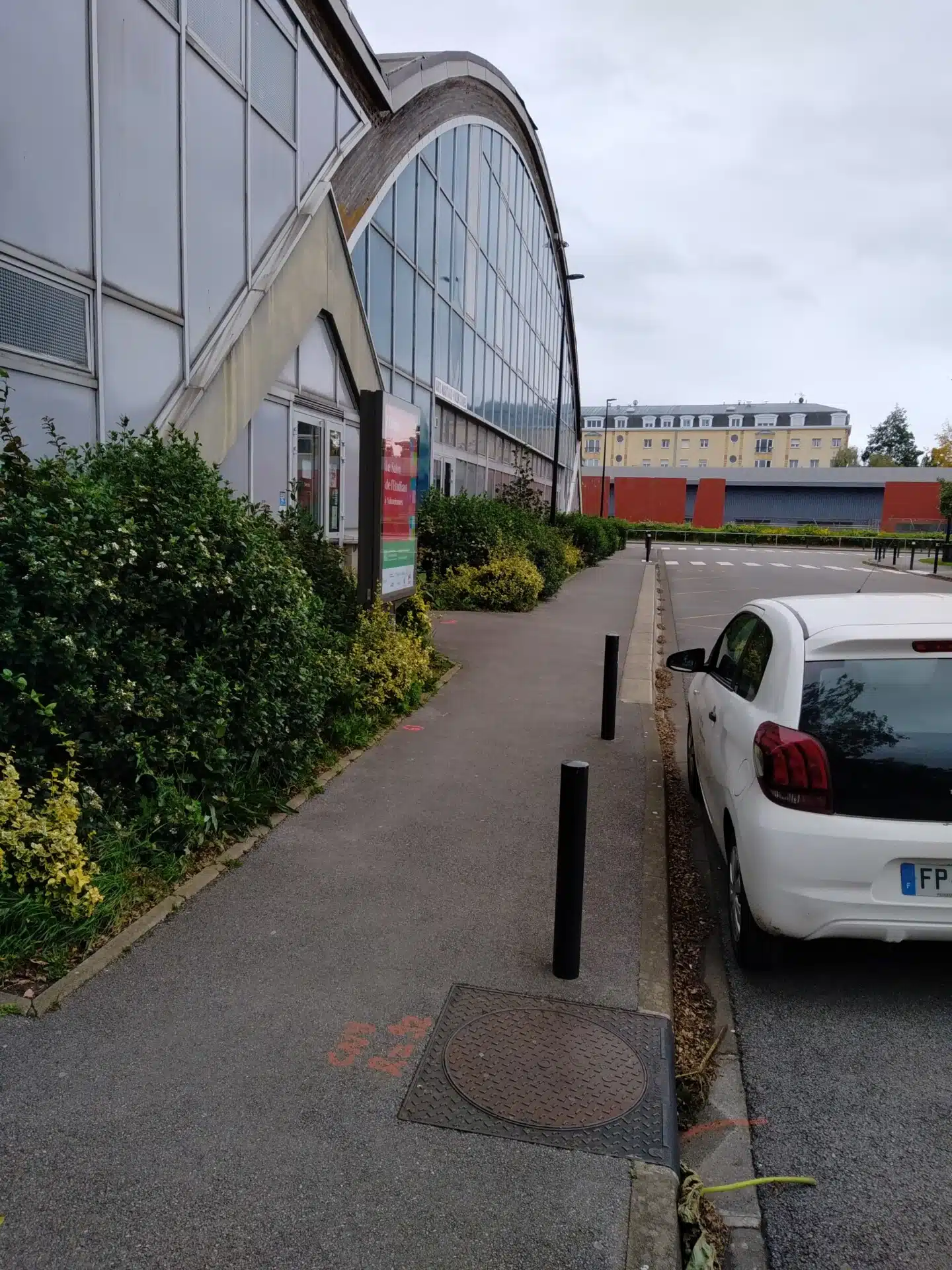 Façade vitrée du bâtiment Pierre Carou, moderne à toit incurvé, bordé d’un trottoir avec des plantations. Une voiture blanche est stationnée sur le côté, ajoutant une touche urbaine à l’environnement.